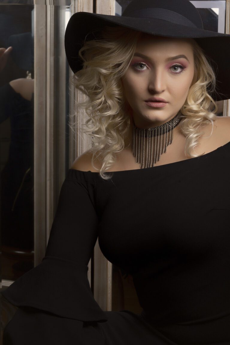 Woman with blonde curly hair wearing a black off-shoulder top and a statement necklace.