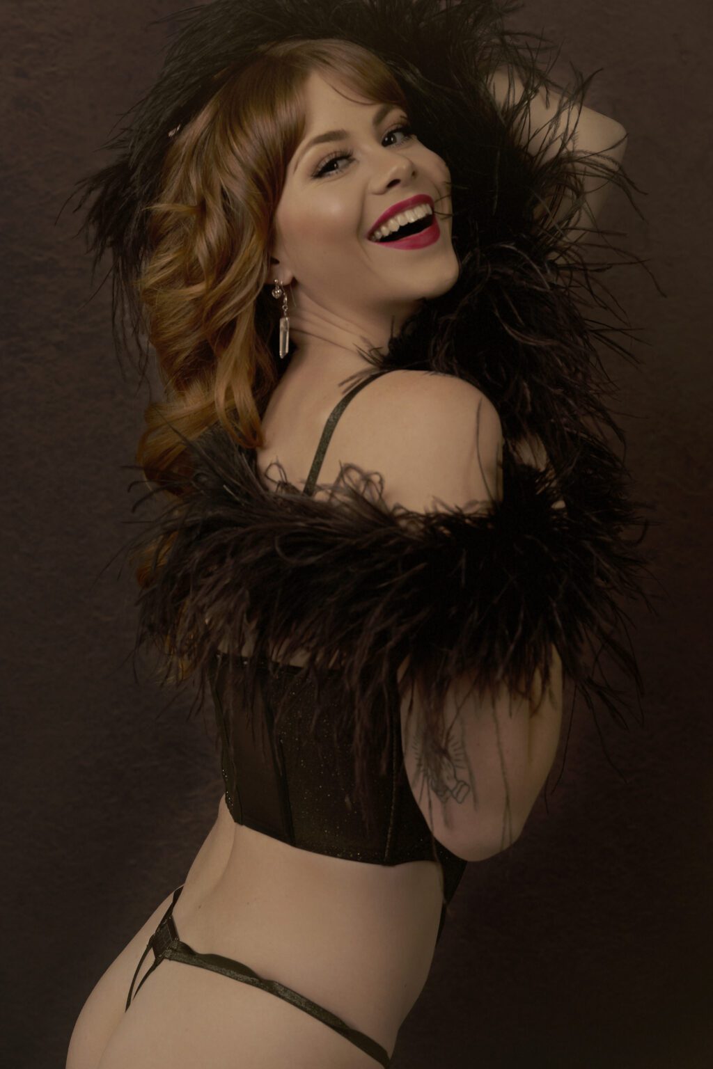 Boudoir portrait of a smiling woman in black lingerie with feather accents, styled in a glamorous vintage-inspired look at Miriam’s Boudoir Studio in Michigan.