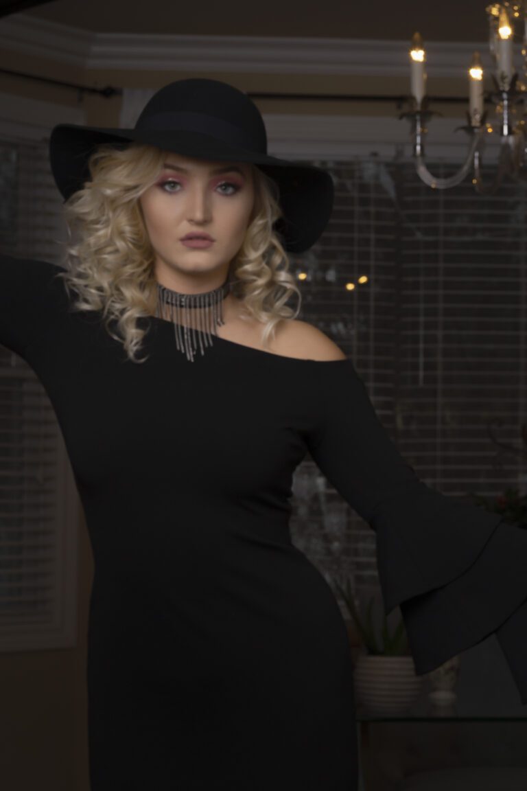 Woman with curly blonde hair in a black off-shoulder dress and hat.